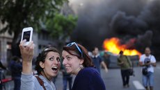 Туристы фотографируются на фоне горящего контейнера во время майской демонстрации в Барселоне, Испания Туристы фотографируются на фоне горящего контейнера во время майской демонстрации в Барселоне, Испания